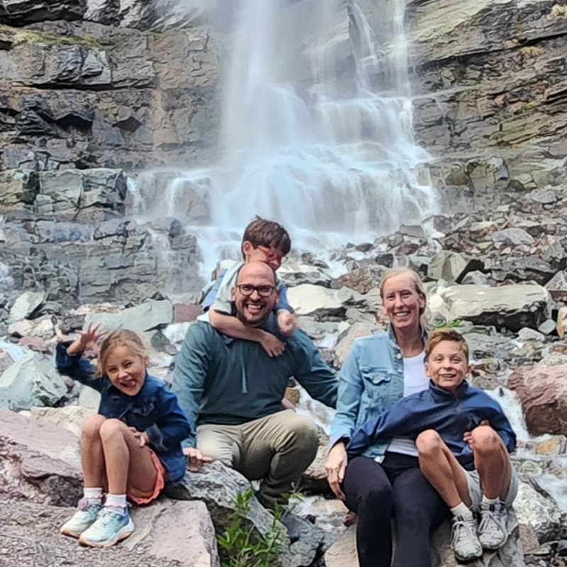 A family of five poses in front of a waterfall surrounded by rocks and greenery.
