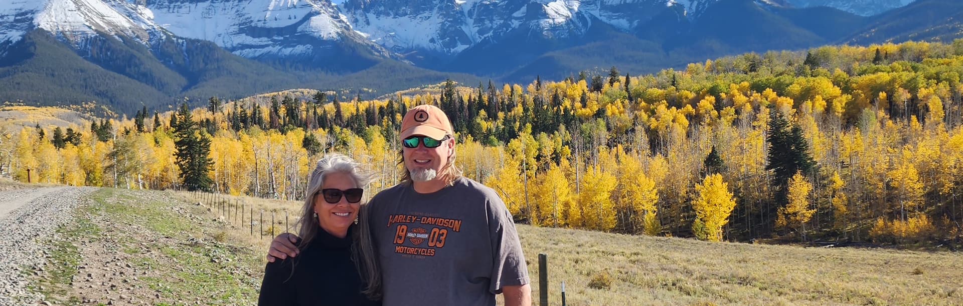 A couple stands together in front of a colorful autumn landscape with mountains in the background.